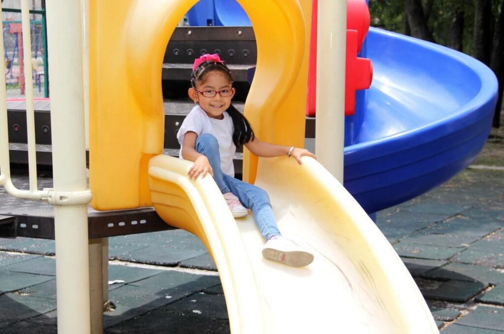 A smiling girl wearing glasses sitting at the top of a slide in the playground.