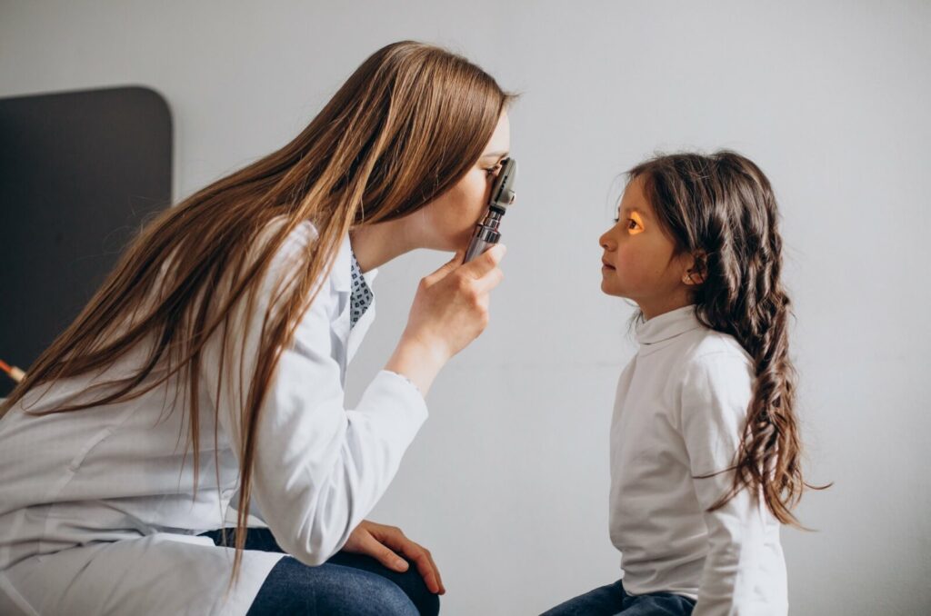 An eye doctor holding an optometric device in her right hand and shining a light into a girls eye during an eye exam.