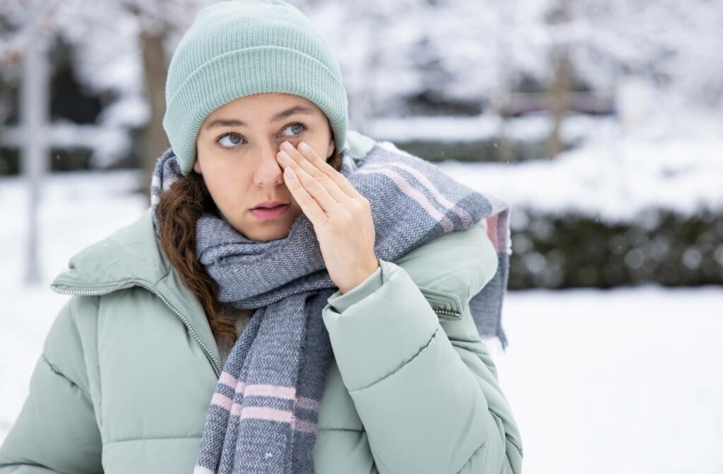 A person rubs their dry eyes while outside in the winter. They are wearing a green winter coat, beanie, and scarf.