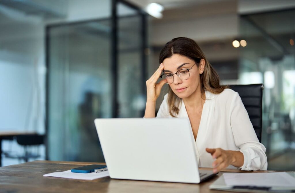 A person wearing glasses trying to work on their computer but struggling with blurry vision and a headache.