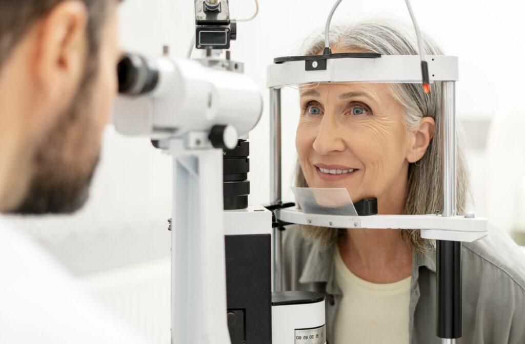 Older woman with gray hair undergoing an eye exam using a slit lamp while an optometrist observes through the microscope.